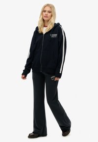 Marineblå zip-up hoodie med hvide sidelister og logo, parret med mørkegrå sweatpants med trykt tekst og en afslappet pasform.