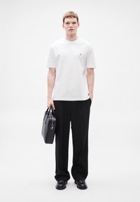 Biele tričko s krátkym rukávom a malým logom, kombinované s čiernymi voľnými nohavicami. Model drží čiernu koženú tašku a má na nohách čierne topánky.