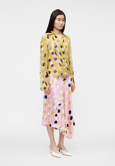 Διάφανη κίτρινη μπλούζα με πολύχρωμες πουά βούλες, σε συνδυασμό με ασύμμετρη ροζ φούστα με παρόμοια σχέδια. Λευκά παπούτσια με μυτερό άκρο.