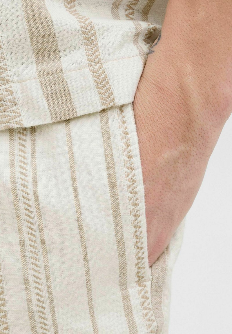 Liinakangas, jossa on beige vaakaraitoja, sivutasku ja koristeompeleet reunassa, lähikuva materiaalin tekstuurista.