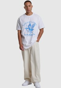 White cotton t-shirt featuring blue graphic print of a cherub with arrows. Paired with loose-fitting cream pants and white sneakers.