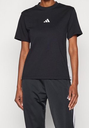 Osoba ubrana w czarną koszulkę Adidas i czarne spodnie dresowe z białymi paskami po bokach, stojąca na jednolitym tle.