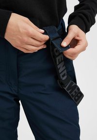 Navy blue outdoor pants with a textured fabric, featuring a black adjustable belt secured with a Velcro strap. Hands fasten the belt.