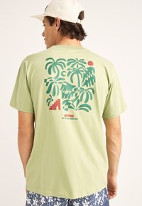 Camiseta ligera verde con un gran gráfico de palmeras, un sol y escaleras en verde oscuro y rojo. Material de algodón suave con mangas cortas.