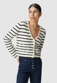 Cardigan in lana a righe nei toni crema e navy, con scollo a V, bottoni perla e due tasche frontali. Indossato con jeans in denim scuri.