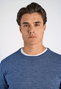 Blauer Sweatshirt mit strukturiertem Strickmuster, Rundhalsausschnitt und gerippten Bündchen, getragen über einem weißen T-Shirt. Einfaches Design, keine sichtbaren Logos.