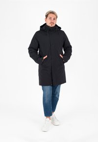 Parka nera con cappuccio, dotata di tasche frontali e chiusura con zip, realizzata in materiale liscio e resistente alle intemperie; indossata sopra jeans blu e sneakers bianche.