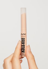 Correcteur Pro Fix Stick de NYX dans un tube cylindrique transparent de couleur pêche. Du texte noir sur le tube présente les détails du produit.