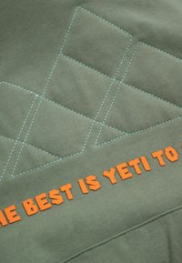 Zielony materiał z niebieskimi przeszyciami tworzy wzór diamentowy. Fraza "NAJLEPSZE JESZCZE PRZED NAMI" jest wyświetlana dużymi pomarańczowymi literami.