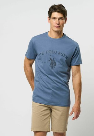 Svijetloplava pamučna majica kratkih rukava s tamnim printom igrača poloa i natpisom "U.S. POLO ASSN" centriranim iznad. U kombinaciji s be�ž kratkim hlačama.