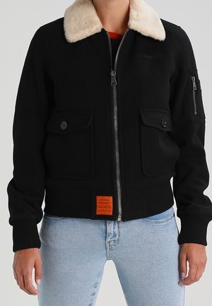 Schwarze Bomberjacke mit beigem Lammfellkragen, Frontreißverschluss, geknöpfte Klappentaschen, Reißverschlusstasche am Ärmel, getragen mit hellblauen Jeans.