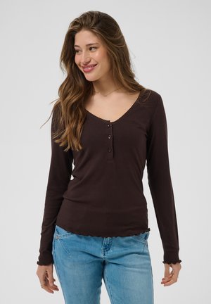 Jeune femme aux longs cheveux bruns, portant un haut marron foncé à manches longues et un jean bleu, souriante et regardant légèrement vers sa gauche.