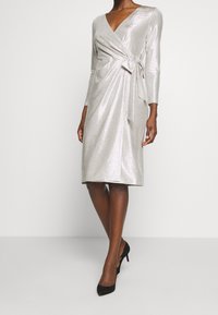 Silverfärgad metallisk omlottklänning med långa ärmar, V-ringning och sidoknytning, med knälång längd och slät textur. Svarta högklackade skor.