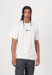 Camiseta de algodón blanca con mangas cortas, cuello redondo y logo de "ellesse" en negro. Lleva puesta con pantalones cargo grises, de pie contra un fondo liso.