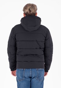 Giacca a vento nera con cappuccio, caratterizzata da cuciture orizzontali, polsini elasticizzati e una lunghezza fino alla vita. Indossata sopra dei jeans blu.