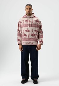 Sweat à capuche crème avec motifs de chevaux bordeaux et cactus. Coupe ample avec une poche kangourou et un cordon de serrage, associé à un pantalon large bleu marine.