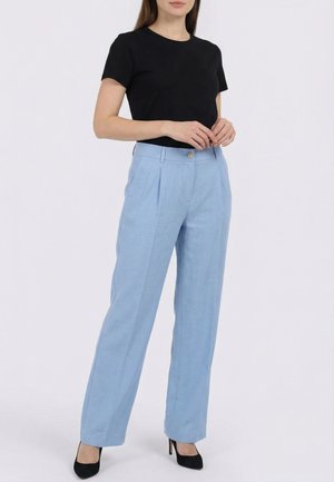 Femme portant un haut noir à manches courtes, un pantalon large taille haute bleu clair et des chaussures à talons noirs, debout devant un fond blanc.