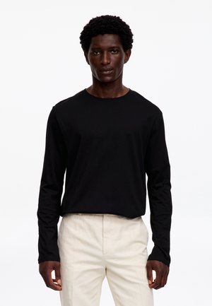 Uomo con capelli ricci scuri che indossa una camicia nera a maniche lunghe e pantaloni beige chiaro, in piedi davanti a uno sfondo bianco uniforme.