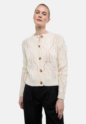 Femme portant un cardigan en tricot couleur crème avec des boutons en bois et un pantalon noir, debout devant un fond clair uni.