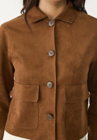 Brown suede jacket with four buttons down the front, two front flap pockets, and a pointed collar worn by a person.