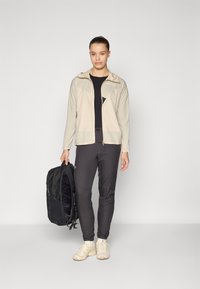 Veste légère beige avec capuche, t-shirt noir, pantalon slim fit foncé, chaussures beige et sac à dos noir. Design simple et look fonctionnel.