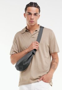Sac en bandoulière en tissu gris texturé, présentant une forme courbée et une sangle ajustable, porté par une personne en polo beige.
