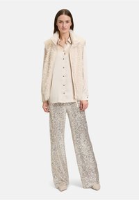 Faux-Fur-Creme-Weste über einem hellen Hemd mit Knöpfen; silberne Pailletten-Hosen mit weitem Bein; Schuhe in neutralen Farbtönen; glatte Texturen und glänzende Oberflächen.