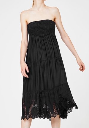 Robe midi noire bustier avec corsage smocké, étages froncés et ourlet orné de dentelle, portée par une personne debout à la peau claire.