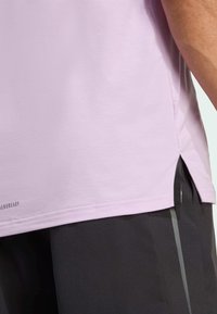 Camisa corta de manga corta en color púrpura claro, hecha de una tela suave, con aberturas laterales y un logo impreso cerca del dobladillo. Combinada con pantalones oscuros de textura.