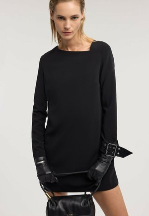 Femme portant une robe noire à manches longues avec des gants et un large détail de boucle, tenant un sac à main en cuir noir devant un fond uni.