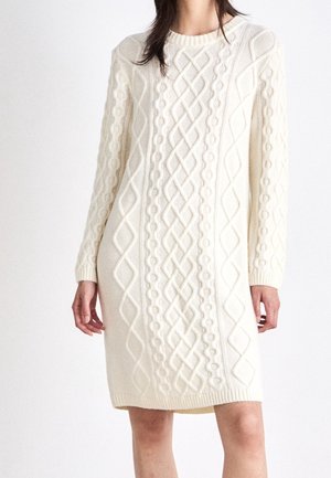 Woman wearing a knee-length cream cable knit sweater dress with long sleeves and a round neckline.