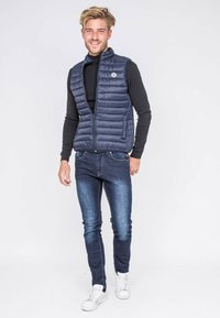 Gilet matelassé bleu marine avec fermeture éclair à l'avant et design matelassé horizontal, porté sur un t-shirt noir à manches longues. Associé à un jean slim et des baskets blanches.