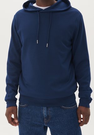Uomo che indossa una felpa con cappuccio blu scuro tinta unita con cordini e jeans blu, in piedi davanti a uno sfondo chiaro.
