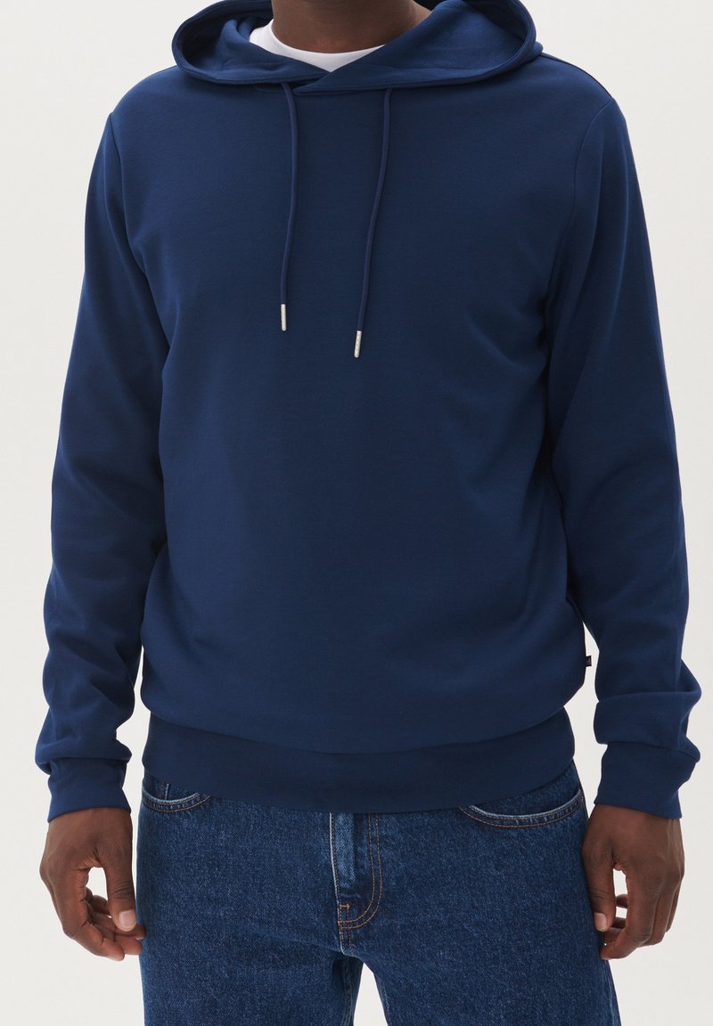 Homme portant un sweat à capuche bleu foncé uni avec cordons et un jean bleu, debout devant un fond clair.