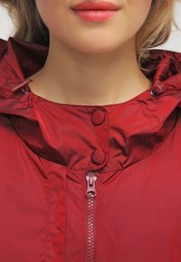 Czerwona lekka kurtka z wysokim kołnierzem, z centralnym zamkiem błyskawicznym i zapięciem na napy, gładką teksturą oraz regulowanymi sznurkami.