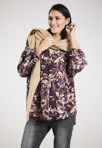 Blumenmuster-Bluse mit langen Ärmeln in tiefburgunderrot und Creme, drapiert mit einem hellbraunen Pullover. Die Bluse hat farbenfrohe botanische Muster und geraffte Bündchen.