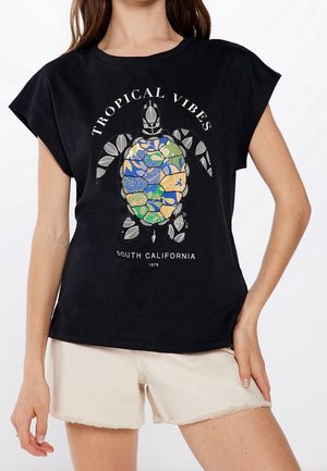 Vrouw draagt zwart T-shirt met kleurrijke schildpadgrafiek en tekst "Tropical Vibes South California 1979", gecombineerd met lichtbeige shorts.