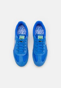 Blå sneakers med en blandning av mocka och mesh-material, platta skosnören, en mönstrad innersula och en grön Puma-logga på tungan.