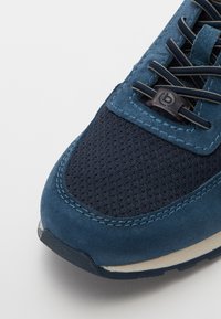 Sapatilha desportiva azul-marinho com parte superior em camurça e painéis em malha, pesponto em contraste e sola em borracha. Destacada com uma aba de cadarço com a marca.