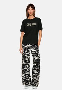 Black t-shirt with "COSMIC" printed in a striped design. Loose-fitting black and white zebra-patterned pants. Silver sneakers.