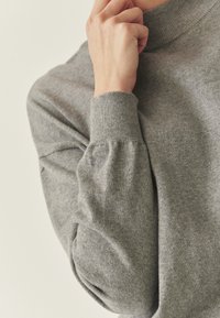 Pull gris tricoté avec un col haut et des manches longues bouffantes, présentant une texture douce et des poignets côtelés.
