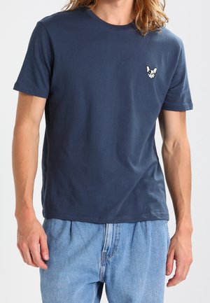Homme portant un t-shirt bleu marine avec un petit logo de bouledogue français blanc sur la poitrine et un jean bleu clair.