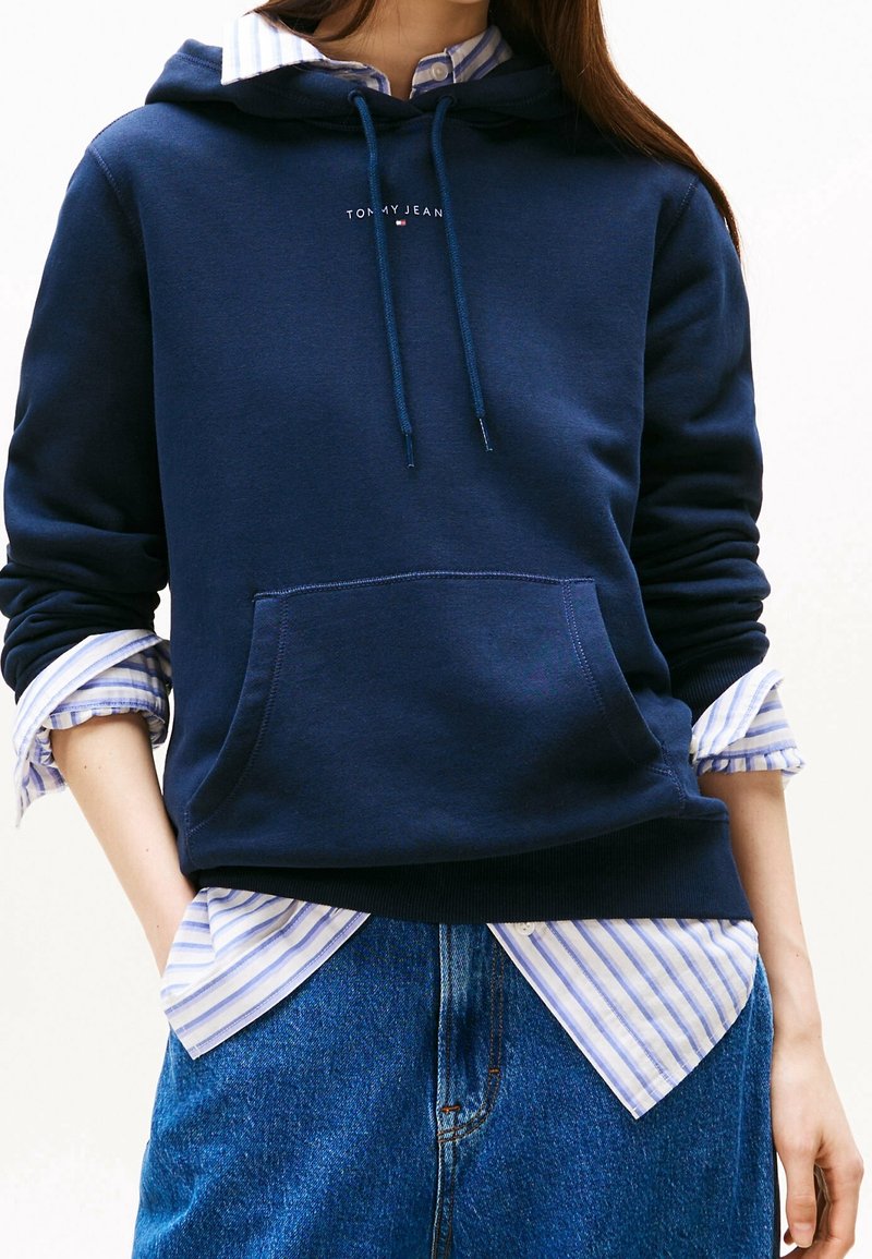 Personne portant un sweat à capuche Tommy Jeans bleu marine sur une chemise à rayures bleues et blanches à boutons avec les manches retroussées et un jean bleu.