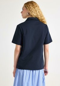 Mørkeblå kortærmet polo shirt med krave, lavet af glat stof. Båret med en lys blå stribet nederdel, set fra bagsiden.