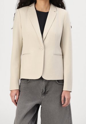 Vrouw draagt een beige blazer met één knoop over een zwart topje en loszittende grijze spijkerbroek, zichtbaar van nek tot heupen op een witte achtergrond.