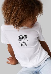Weißes Baumwoll-T-Shirt mit einem schwarzen Druck von "CALVIN KLEIN 1978." Das Design umfasst kurze Ärmel und einen Rundhalsausschnitt.