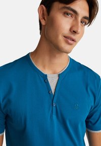 Kratší rukávové tričko typu henley vyrobené z bavlny, s tyrkysovou přední částí, šedým kulatým výstřihem, třemi knoflíky na přední části a logem na hrudi.