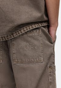 Pantalons cargo marron en tissu résistant, dotés d'une grande poche arrière avec logo en relief et de coutures renforcées le long de la fermeture.
