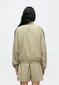 Svetlobéžová bunda s čiernymi bočnými pruhmi, elastickým pásom a vysokým golierom. Na hornej časti chrbta má logo a dlhé rukávy.