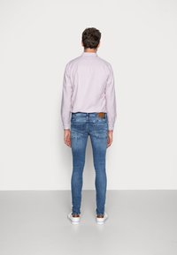Un hombre está de pie frente a una pared gris claro uniforme, vistiendo una camisa de manga larga rosa pálido, jeans ajustados azules y zapatillas blancas con detalles marrones.